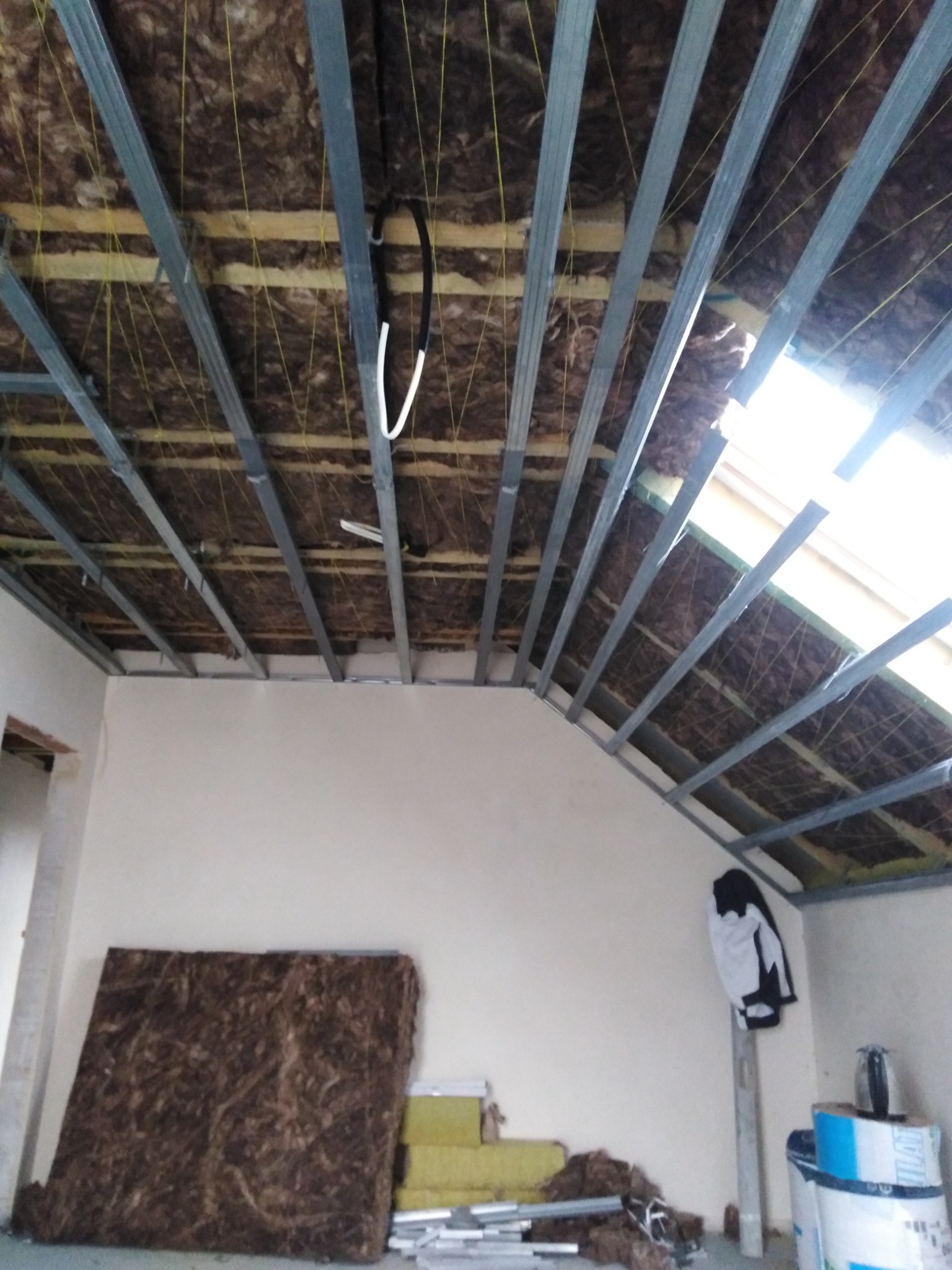 Poddasze w trakcie adaptacji: widoczne metalowe profile konstrukcyjne, izolacja z wełny mineralnej, fragment okna dachowego i przygotowane materiały budowlane przy ścianie.