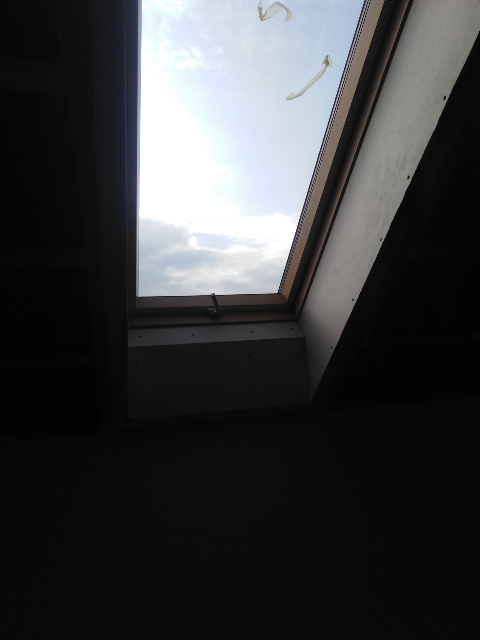 Widok z wnętrza poddasza na okno dachowe, z widoczną płytą karton gips na skosie dachu i fragmentami taśmy malarskiej na szybie okna.