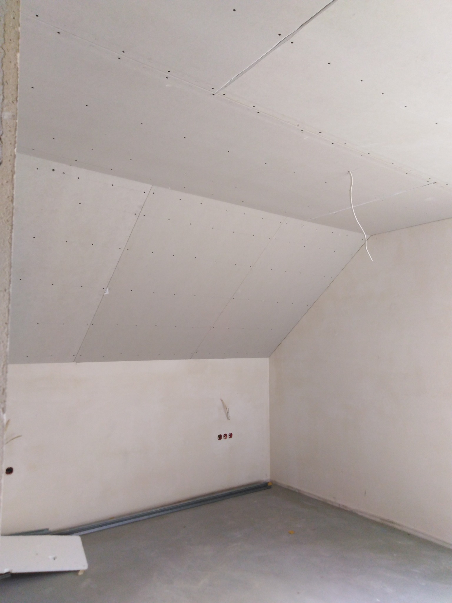 Pokój w trakcie wykańczania, widoczne płyty kartonowo-gipsowe na skośnym suficie poddasza, ściany przygotowane do dalszych prac, wystające przewody elektryczne i profile podłogowe.