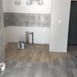 Wnętrze w trakcie remontu: częściowo ułożona podłoga z paneli imitujących drewno, widoczne narzędzia, instalacja wodna i fragment ściany z szarą fakturą betonu.