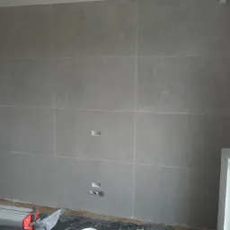 Ściana wyłożona jasnoszarymi płytami wielkoformatowymi z widocznymi fugami, z przygotowanymi otworami na gniazdka elektryczne i puszkę instalacyjną, grzejnik przy ścianie, narzędzia glazurnicze...