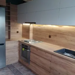 Nowoczesna kuchnia z drewnianymi frontami, szarymi szafkami g&oacute;rnymi i blatem z wbudowaną płytą grzewczą oraz czarnym zlewozmywakiem, oświetlona listwami LED pod szafkami.