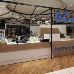 Stoisko z hummusem w centrum handlowym, z drewnianym blatem i niebieskim napisem 'hummus', menu nad głową i metalową konstrukcją nad stoiskiem.