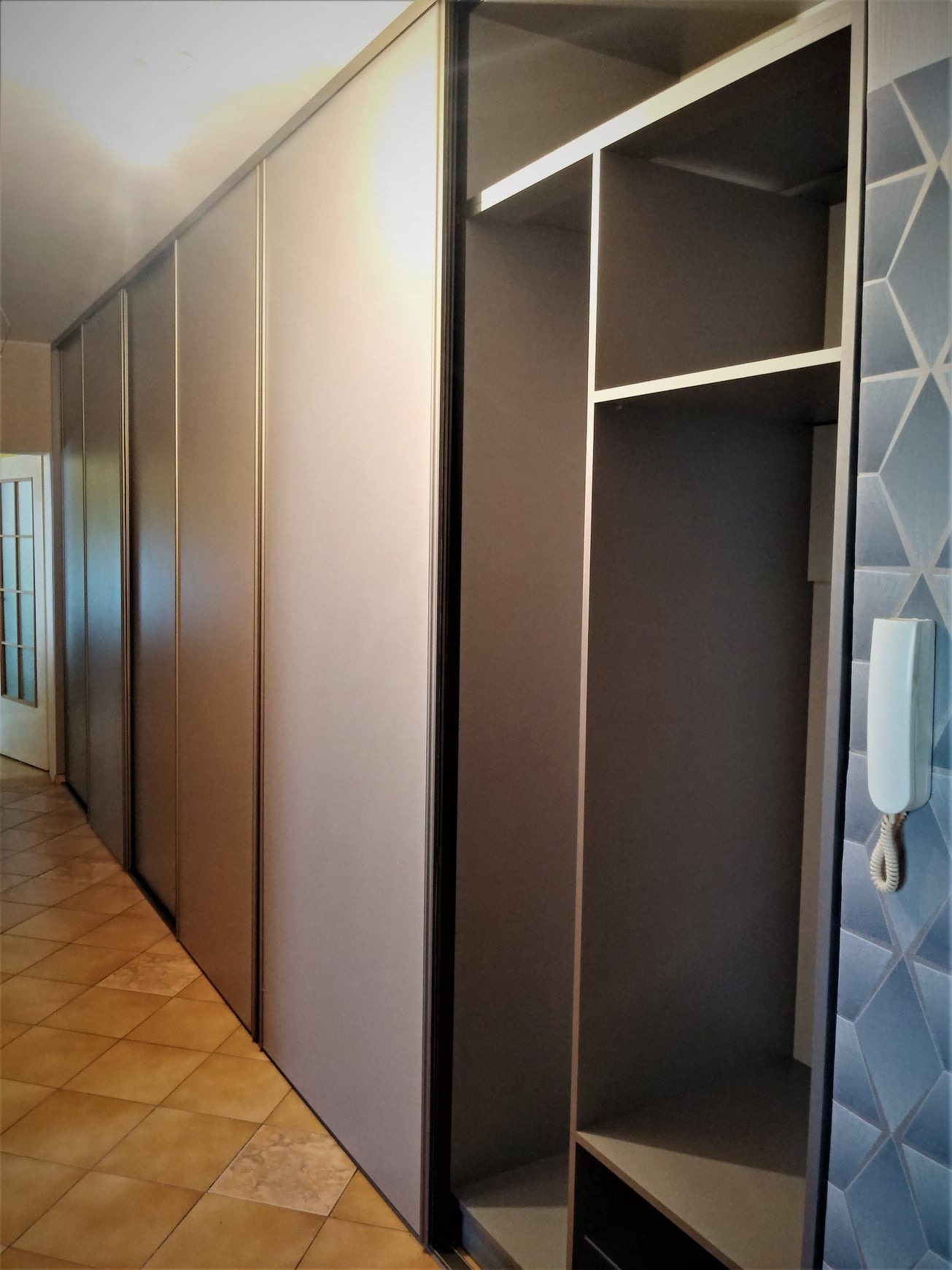 Długa szafa wnękowa z przesuwnymi drzwiami w odcieniach szarości, widoczna otwarta część z półkami, na ścianie dekoracyjna tapeta w geometryczne wzory i domofon.