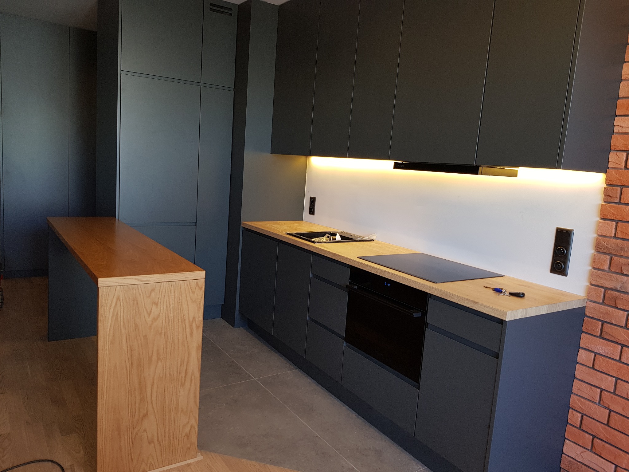 Nowoczesna kuchnia z ciemnoszarymi szafkami i blatem z jasnego drewna, oświetlenie LED pod szafkami, ceglana ściana w tle, klucze leżące na blacie.