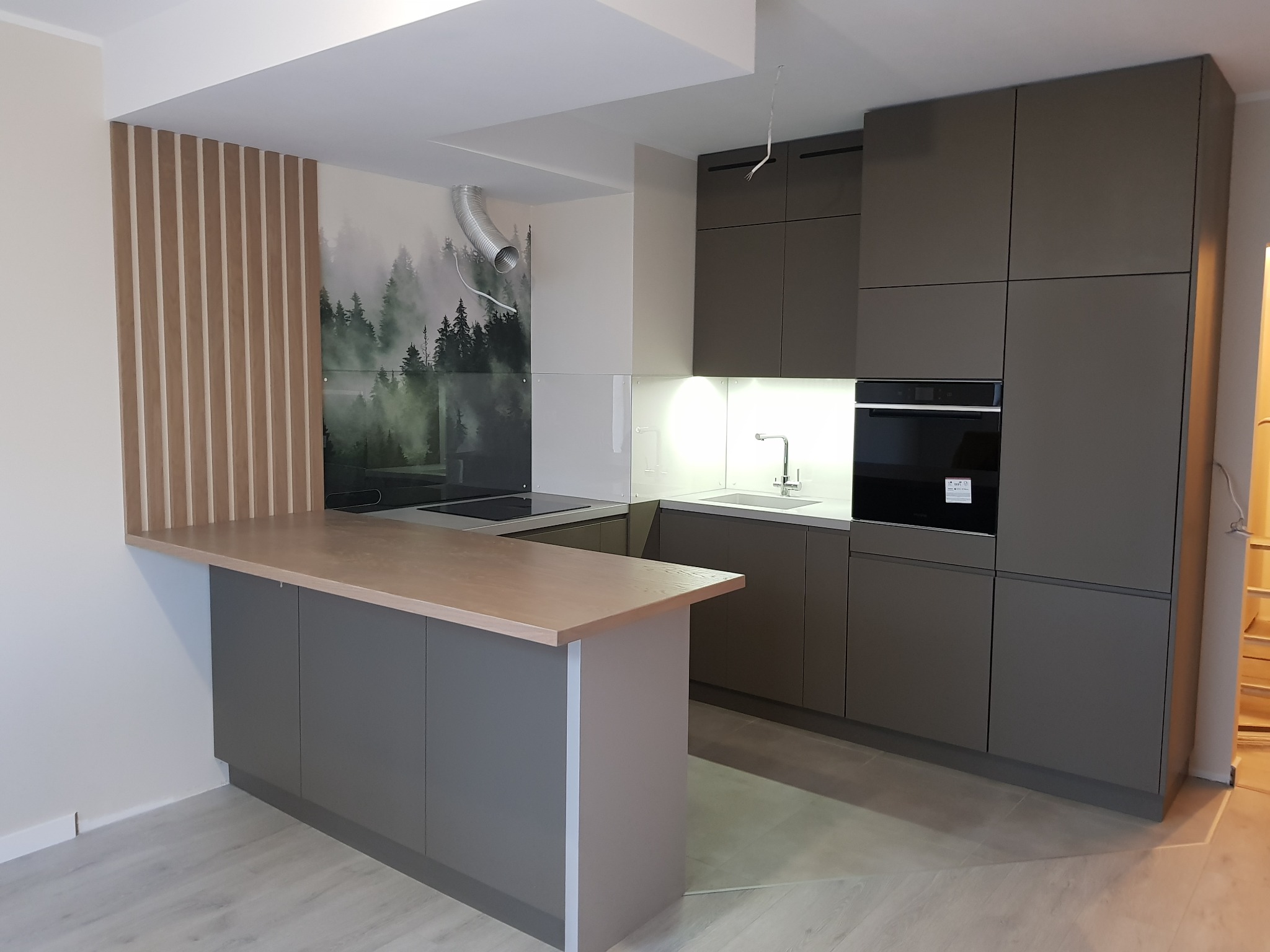Nowoczesna kuchnia z szarymi szafkami, drewnianym blatem i panelem dekoracyjnym z pionowych listew, z motywem leśnym na szklanej ścianie nad blatem.