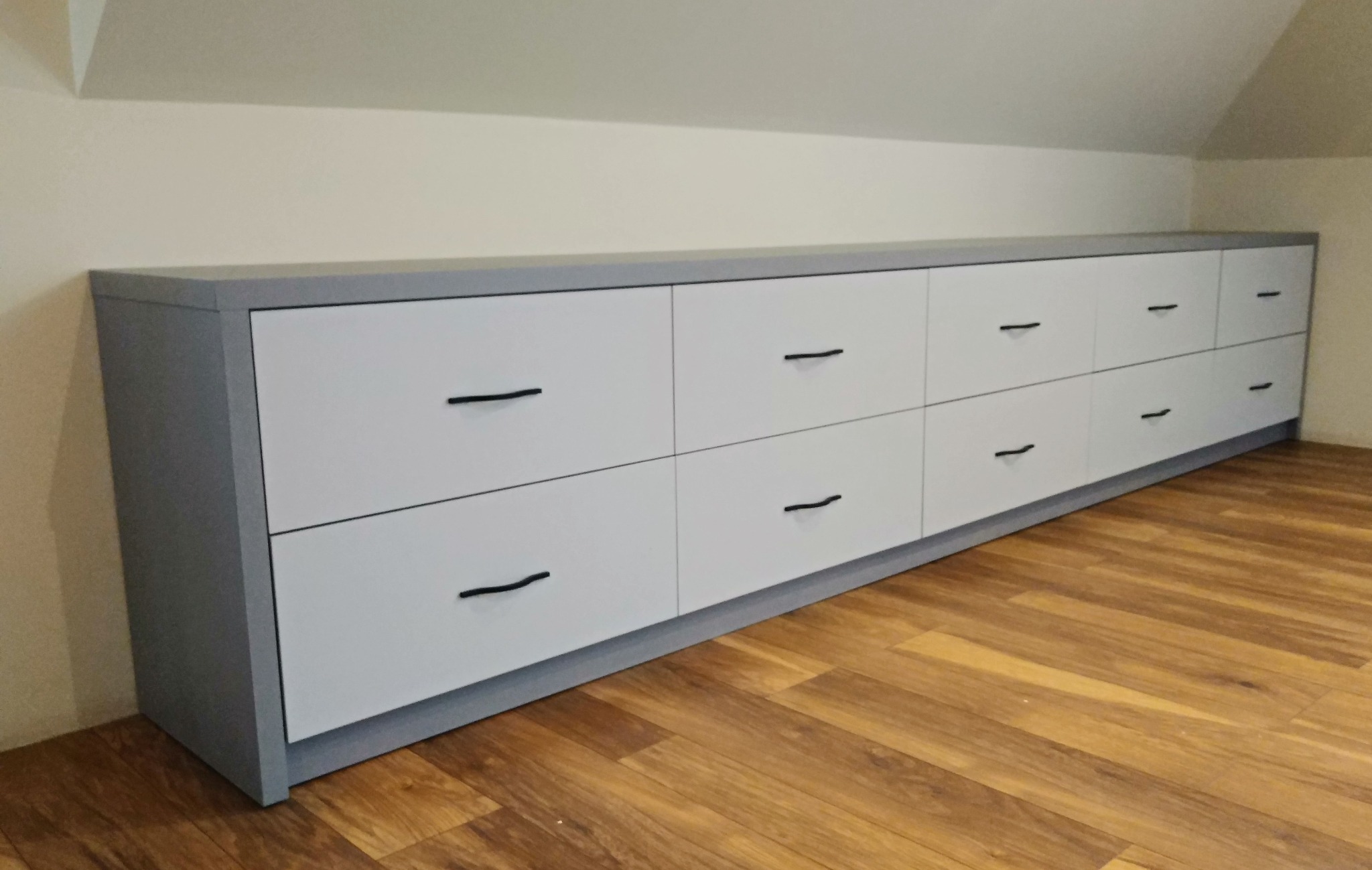 Długa, niska szafka z szufladami w kolorze szaro-białym, z czarnymi uchwytami, umieszczona na drewnianej podłodze pod skosem dachu.