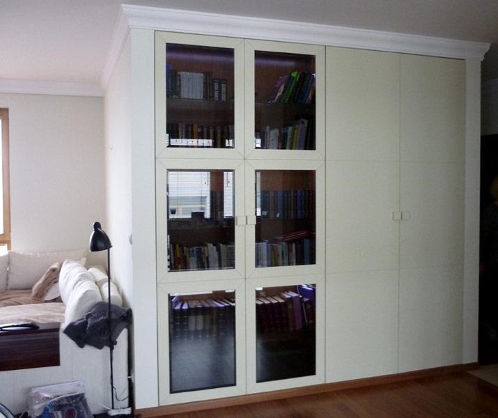 Duża, kremowa szafa wnękowa z przeszklonymi segmentami, wypełniona książkami, w salonie z drewnianą podłogą i narożną sofą.