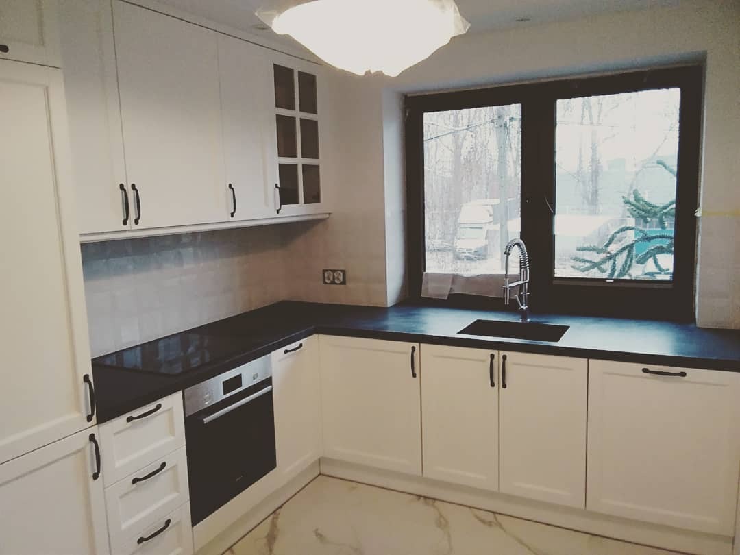 Jasna kuchnia z czarnym blatem i ciemną ramą okienną, widok na zewnątrz w pochmurny dzień.