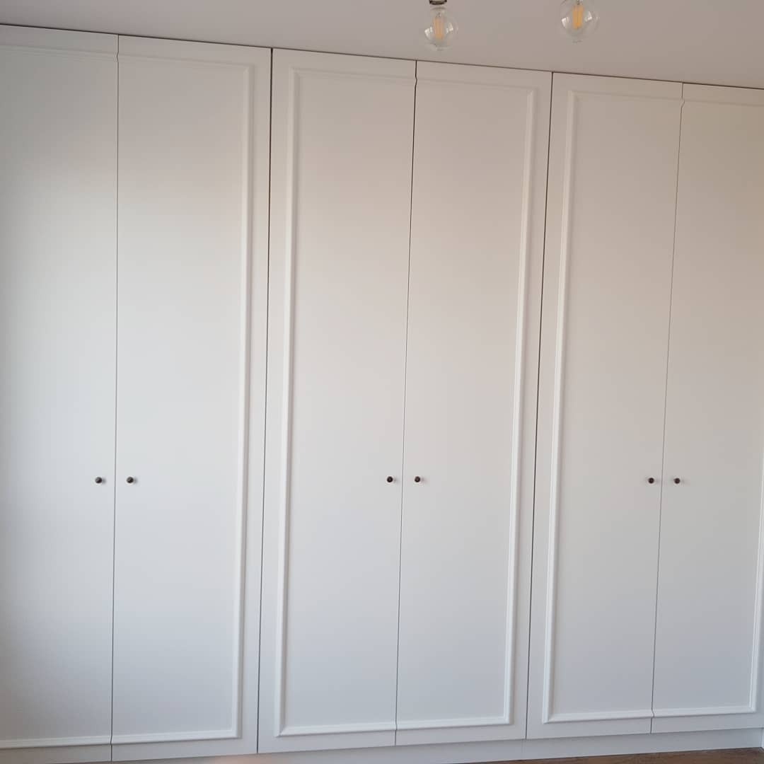 Biała szafa wnękowa z trzema segmentami, każdy z dwoma drzwiami, z delikatnymi ramkami i małymi, okrągłymi uchwytami, oświetlona przez dwie wiszące lampy z żarówkami Edison.