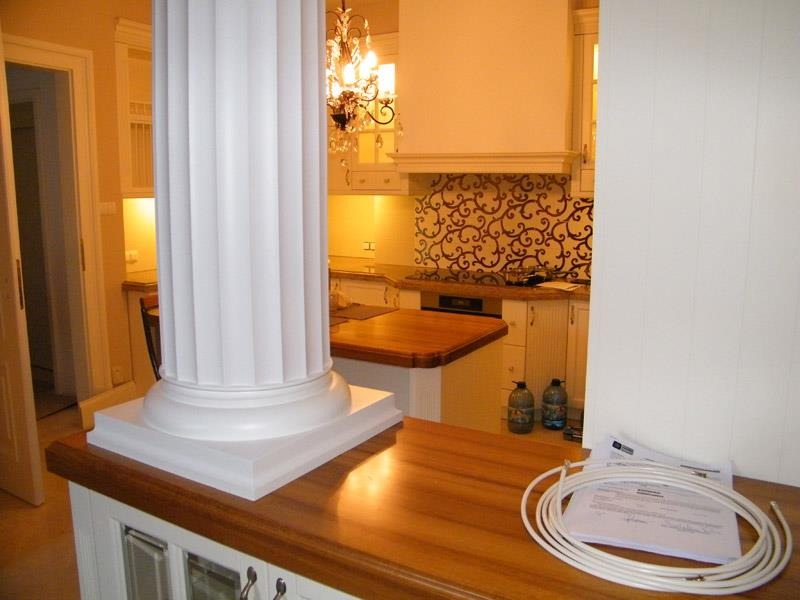 Klasyczna kuchnia z wyspą, drewnianym blatem i dekoracyjną kolumną. W tle wzorzysta ściana nad blatem roboczym i kryształowy żyrandol. Na blacie leżą dokumenty i zwinięty kabel.