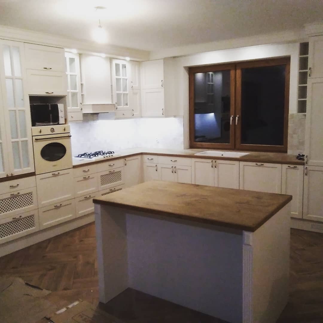 Jasna kuchnia z drewnianym blatem i wyspą kuchenną, parkiet w jodełkę na podłodze, okno w drewnianej ramie, oświetlenie punktowe pod szafkami.