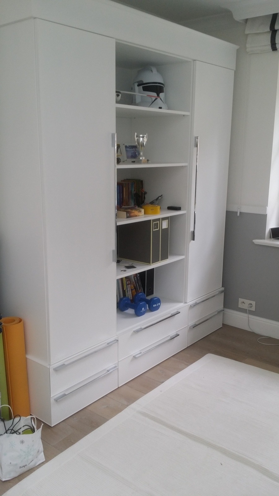 Biała szafa przesuwna z półkami i szufladami, na jednej z półek hełm Star Wars, na dolnych półkach książki i hantle, obok szafy mata do ćwiczeń.