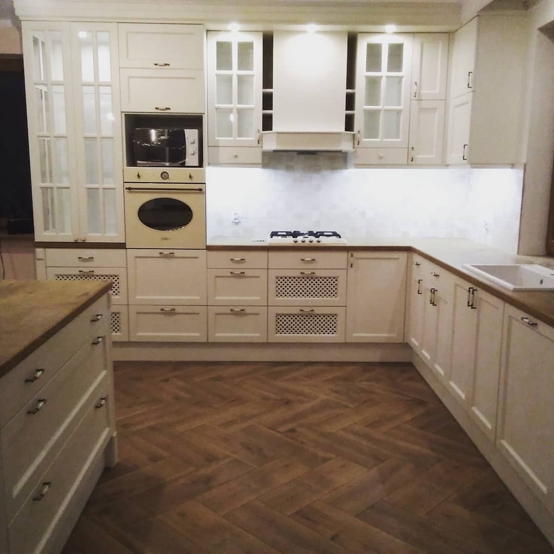 Jasna, klasyczna kuchnia z drewnianym parkietem ułożonym w jodełkę. Zabudowa kuchenna z frezowanymi frontami, szklanymi witrynami i sprzętem AGD w stylu retro. Blat drewniany.