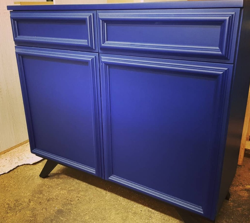 Niebieska szafka z dwiema szufladami i dwojgiem drzwiczek, stylizowana z frezowanymi obramowaniami, ustawiona na niskich, skośnych nóżkach na tle surowej podłogi.