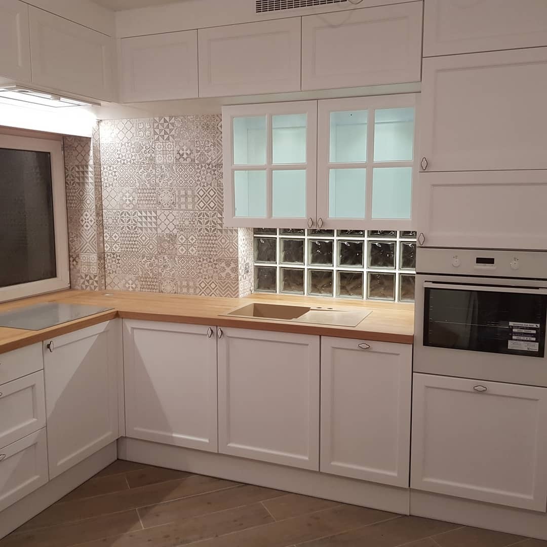 Jasna kuchnia z białymi frontami szafek, drewnianym blatem i dekoracyjnymi płytkami na ścianie, oświetlona blokami z luksferów.