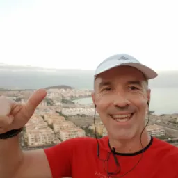 Uśmiechnięty mężczyzna w czerwonej koszulce i czapce z daszkiem, robiący selfie z kciukiem w górę na tle panoramy miasta i morza o zachodzie słońca, z słuchawkami na szyi.