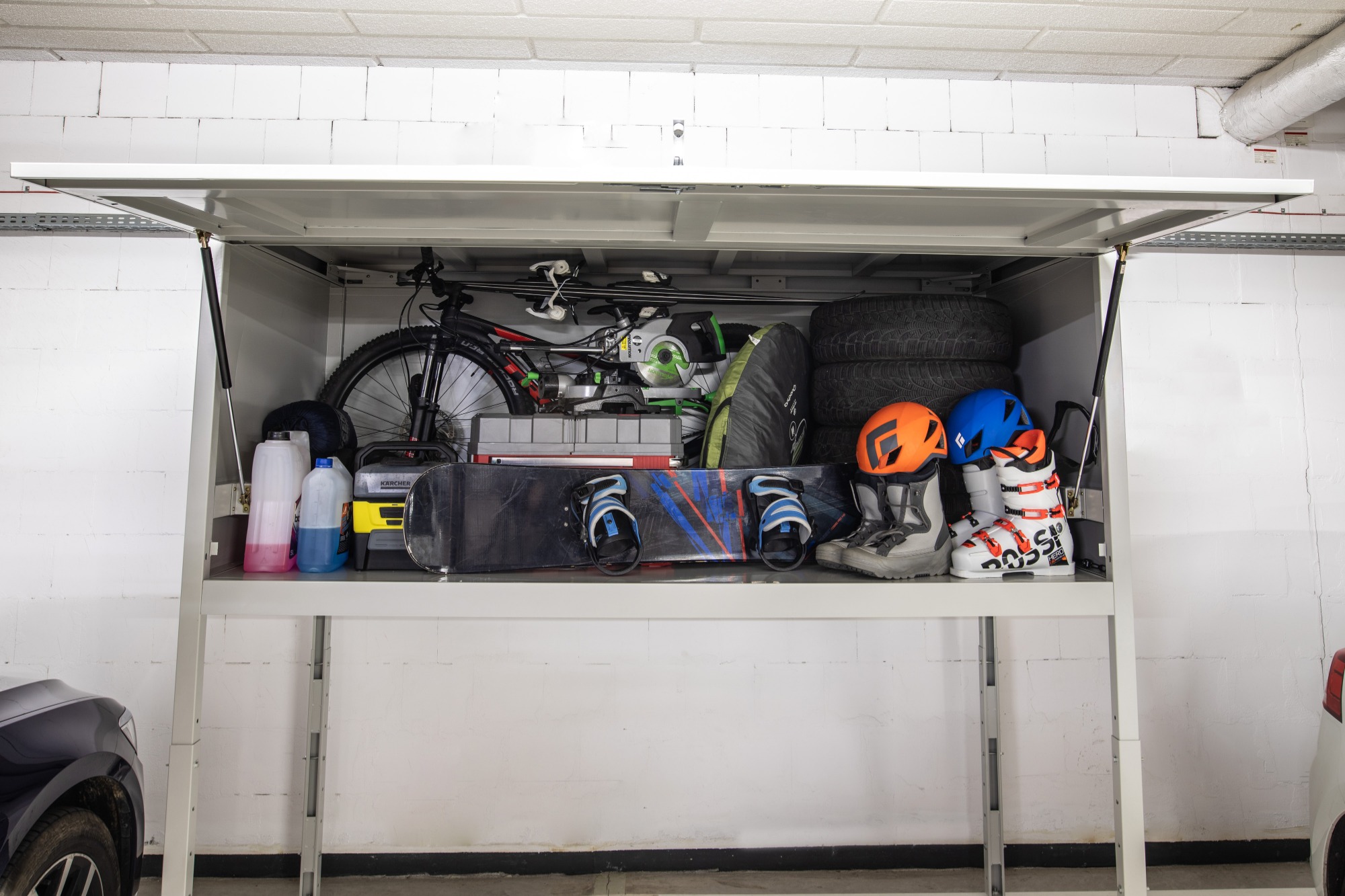 Garażowa szafa z podnoszonymi drzwiami wypełniona rowerem, nartami, snowboardem, oponami, butami narciarskimi i kaskami, z widocznym fragmentem samochodu.