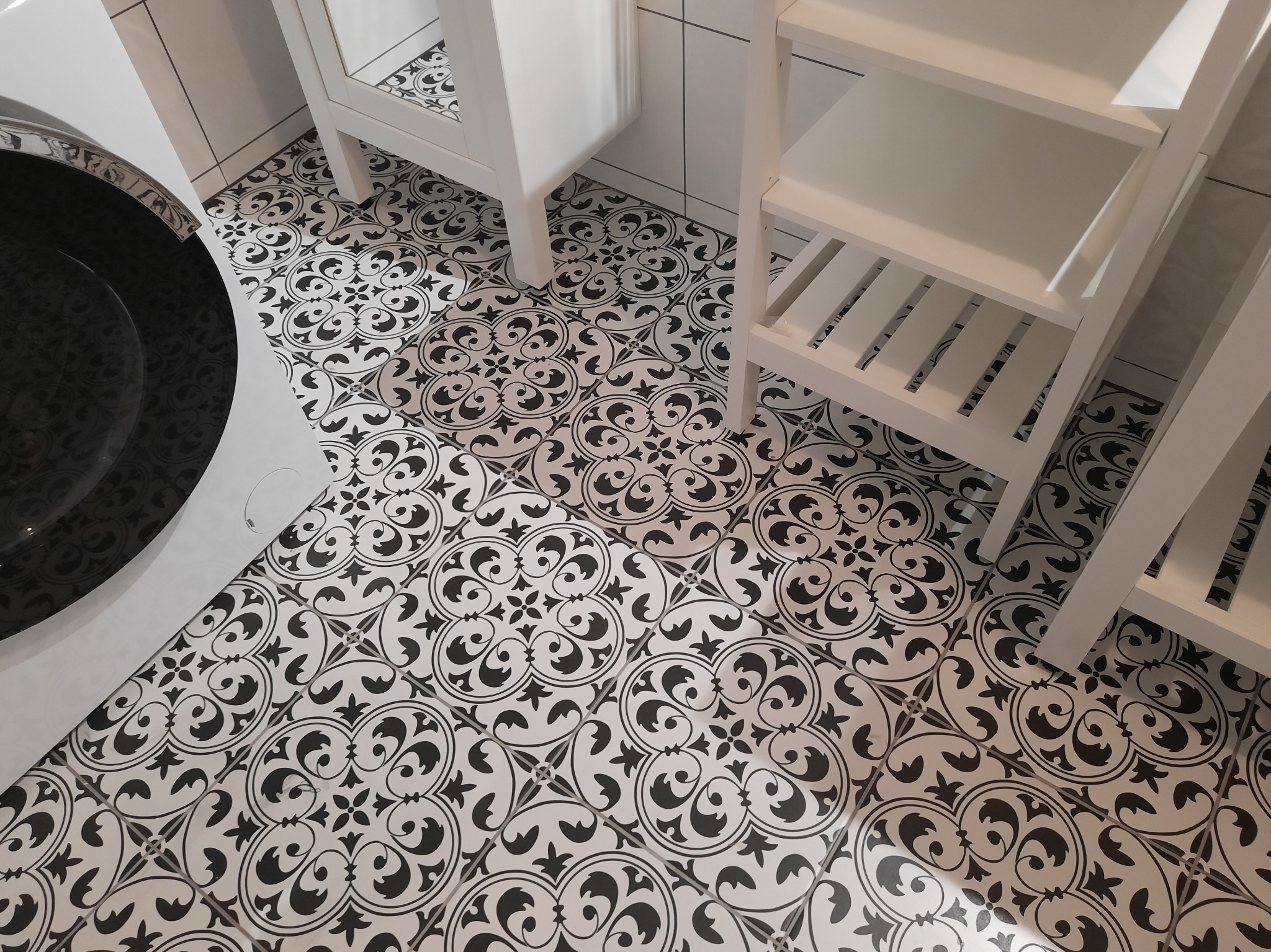 Czarno-białe płytki podłogowe z motywem ornamentów w łazience, widoczne fragmenty umywalki, szafki i regału.