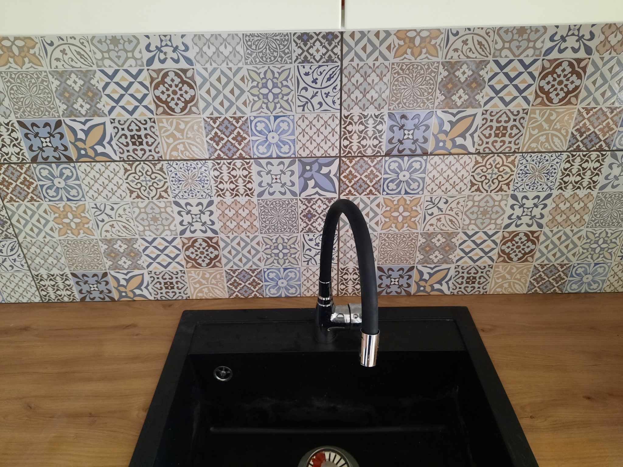 Ściana nad blatem kuchennym wyłożona dekoracyjnymi, wzorzystymi płytkami ceramicznymi w odcieniach beżu, brązu i błękitu, z czarnym zlewem i nowoczesną baterią.