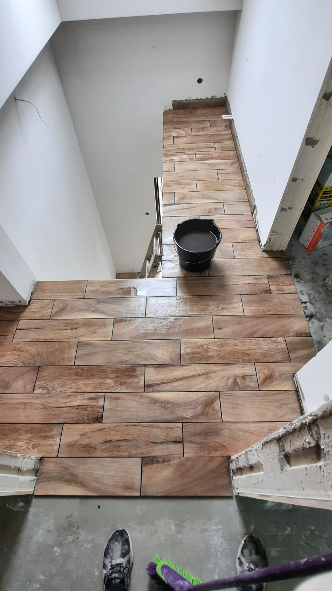 Montaż płytek imitujących drewno na schodach i podłodze, widoczne wiadro z wodą i szczotka do czyszczenia fug.