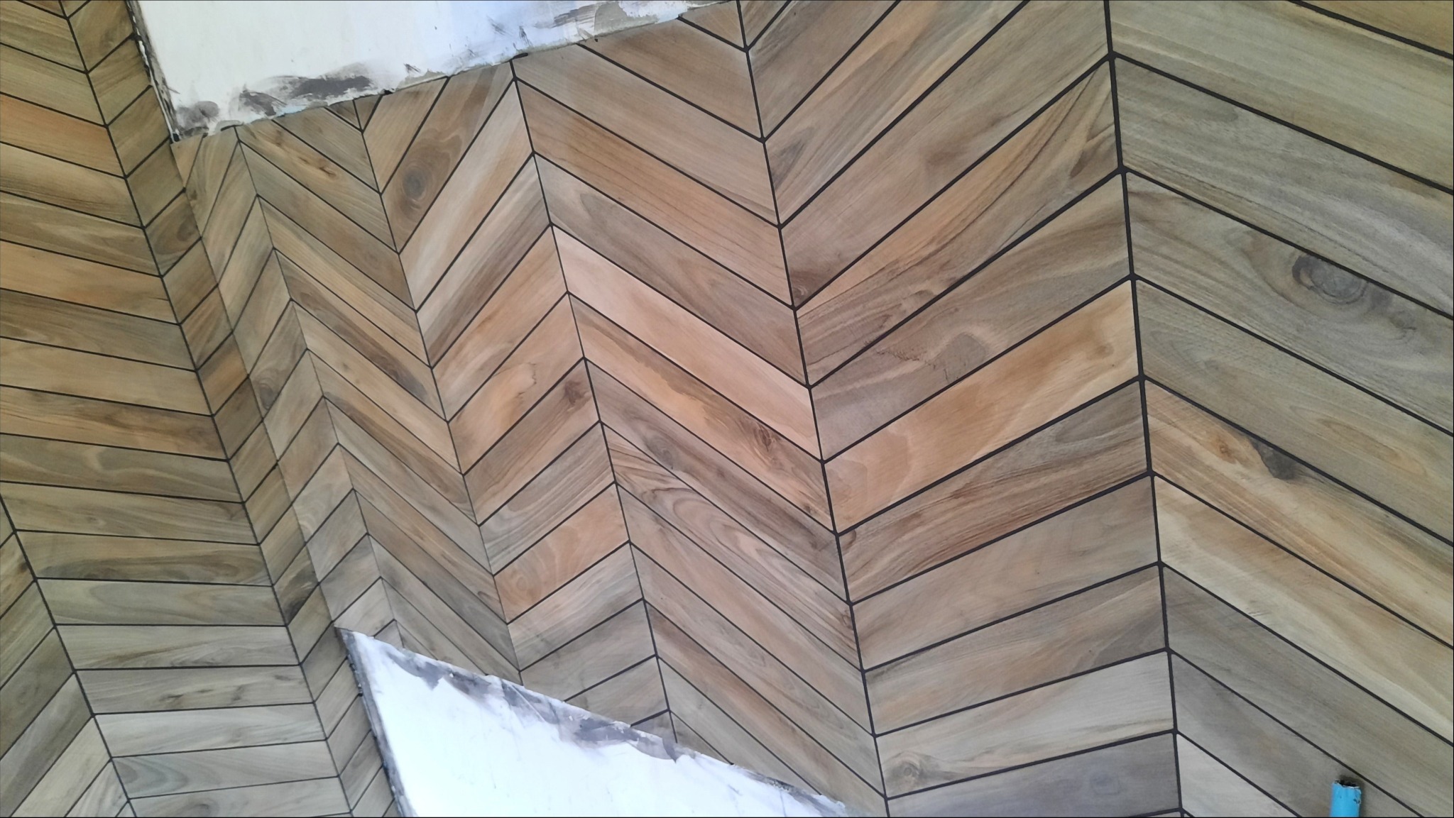 Ułożone w jodełkę drewnopodobne kafelki z ciemnymi fugami na ścianie, widoczne ślady świeżego montażu przy suficie.