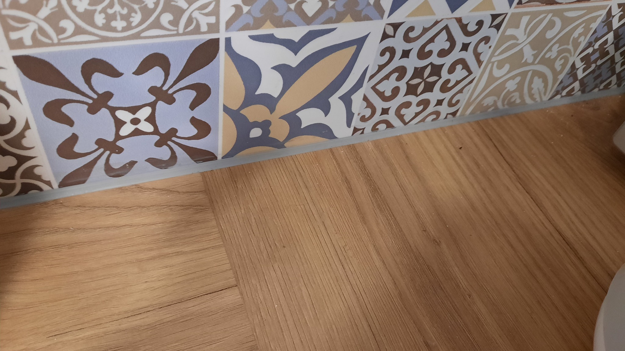 Fragment ściany wykończonej dekoracyjnymi, wzorzystymi kafelkami, stykającej się z drewnianą podłogą.