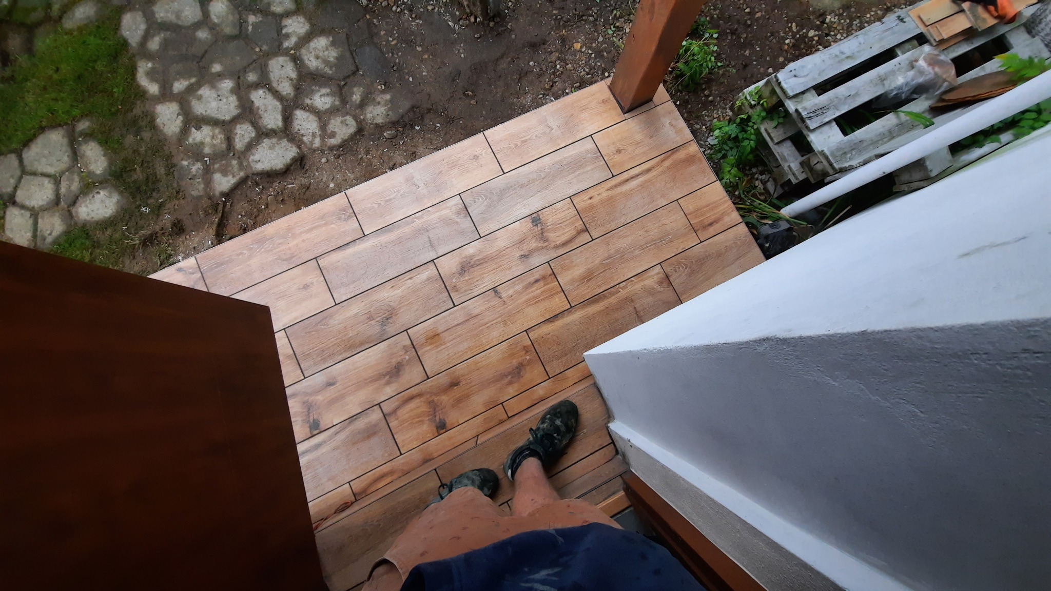 Ułożone kafelki imitujące drewno na zewnątrz, widok z góry na fragment tarasu z widocznymi butami osoby fotografującej, obok biała ściana i ziemia z roślinnością.