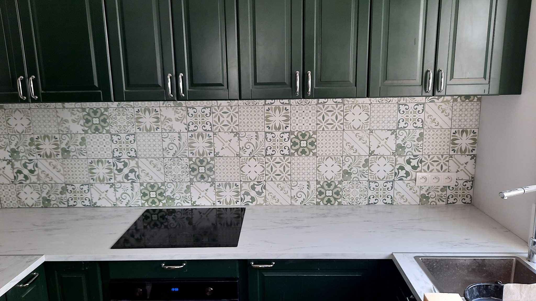 Kuchnia z ciemnozielonymi szafkami, marmurowym blatem i ozdobnymi, zielono-białymi płytkami na ścianie nad blatem.