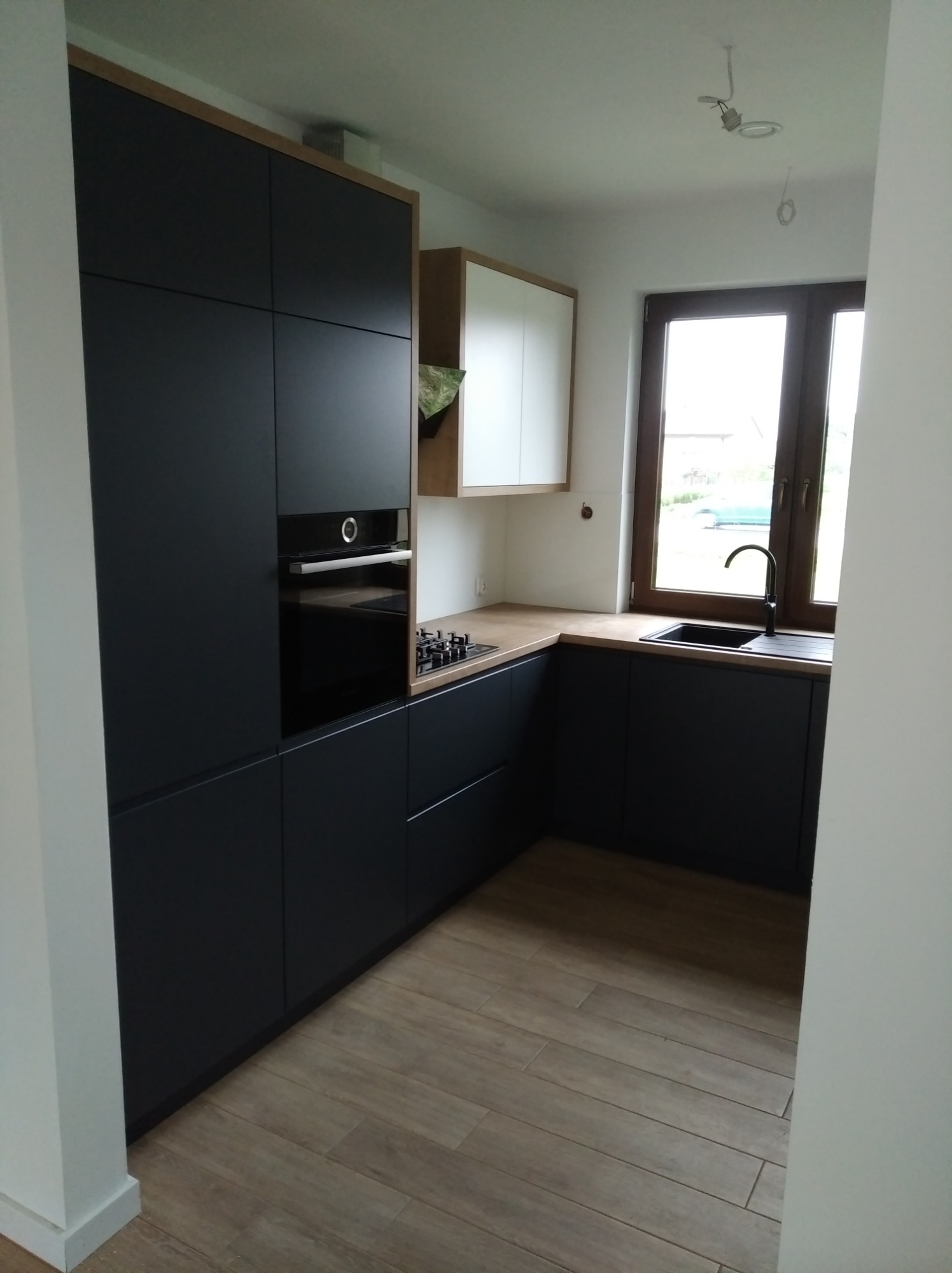 Nowoczesna kuchnia z czarnymi szafkami, drewnianym blatem i podłogą, widok na okno z czarną ramą i zlewozmywak, oświetlenie sufitowe w trakcie montażu.