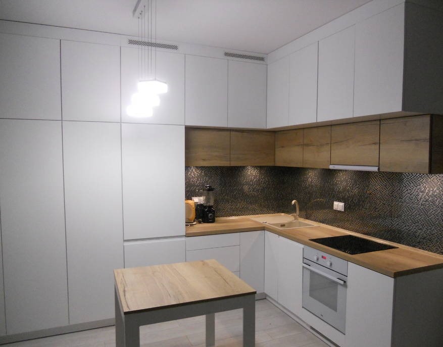 Nowoczesna kuchnia w bieli i drewnie z geometrycznym wzorem na ścianie nad blatem, oświetlona wiszącą lampą z trzema kwadratowymi kloszami.