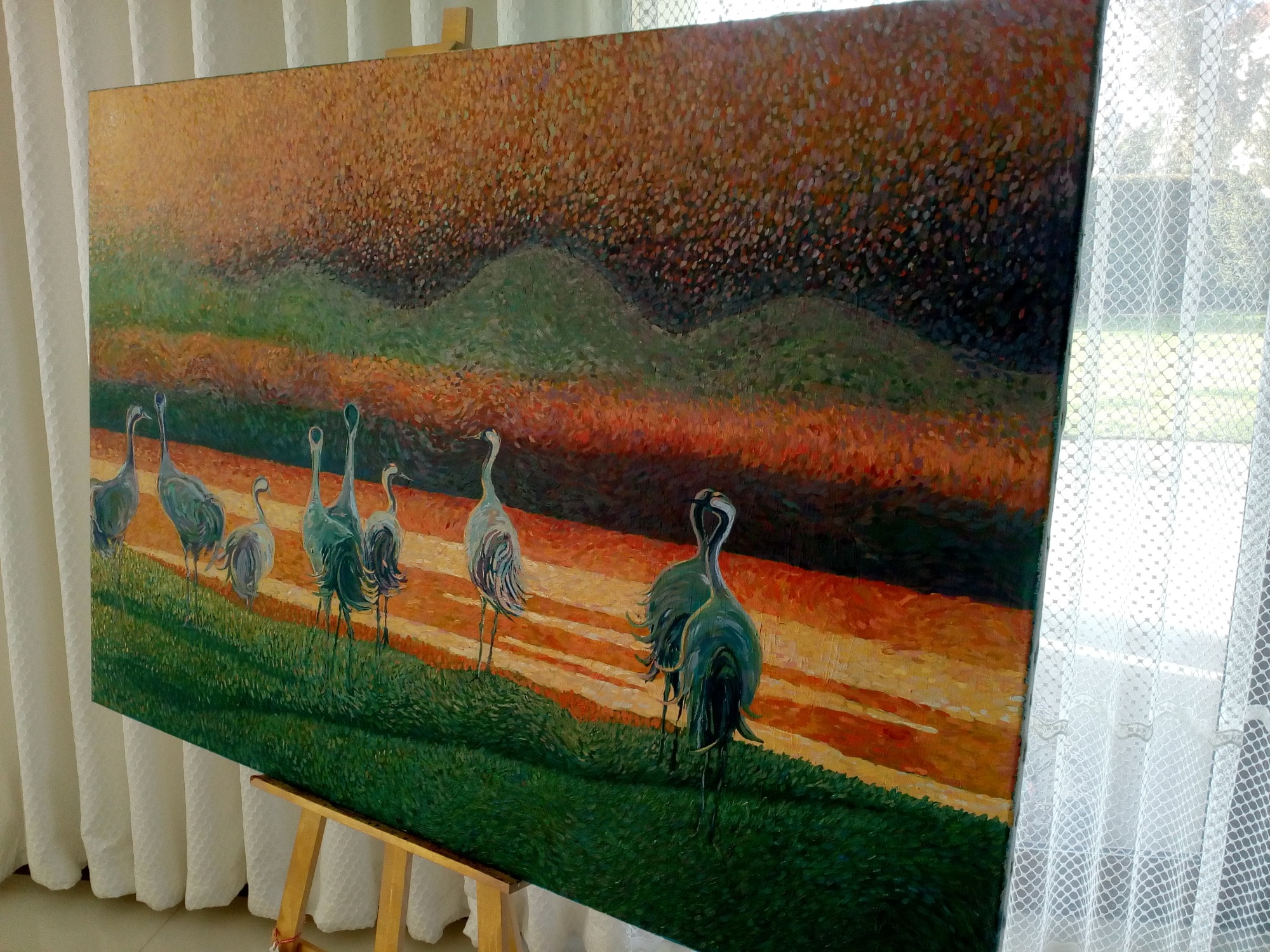 Obraz olejny przedstawiający stado żurawi na tle zachodzącego słońca, z widoczną fakturą farby i subtelnymi przejściami kolorystycznymi, ustawiony na drewnianej sztaludze przy oknie z białą...