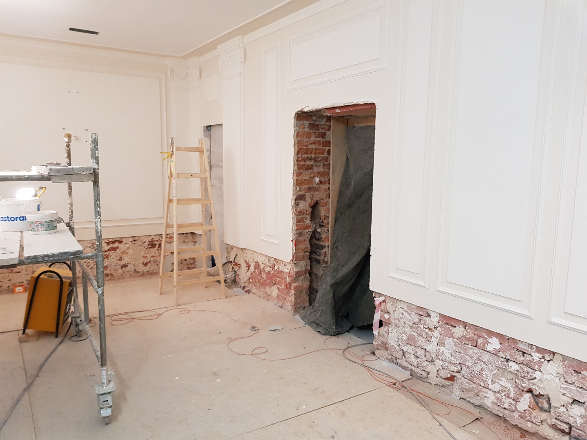 Pokój w trakcie prac wykończeniowych: białe panele ścienne z dekoracyjnymi listwami, odsłonięta cegła poniżej, drabina oparta o ścianę, rusztowanie z wiadrami farby i narzędziami, przygotowane...