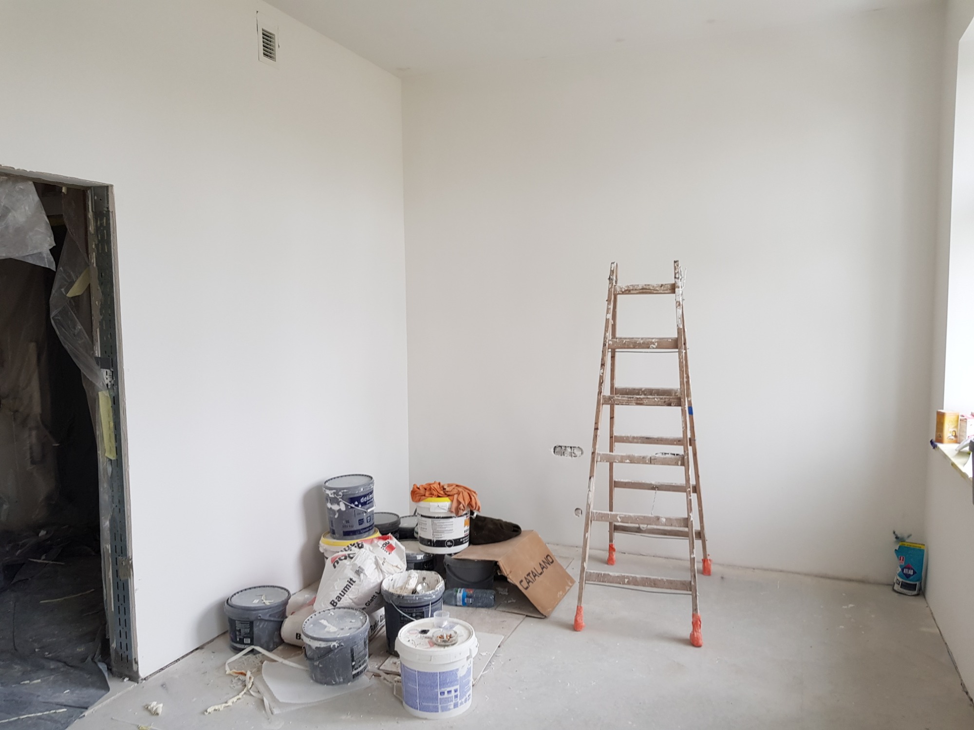 Pokój w trakcie prac wykończeniowych: świeżo pomalowane białe ściany, drewniana drabina oparta o ścianę, wiaderka z farbami i worki z zaprawą budowlaną zgromadzone w rogu, zabezpieczone folią drzwi...