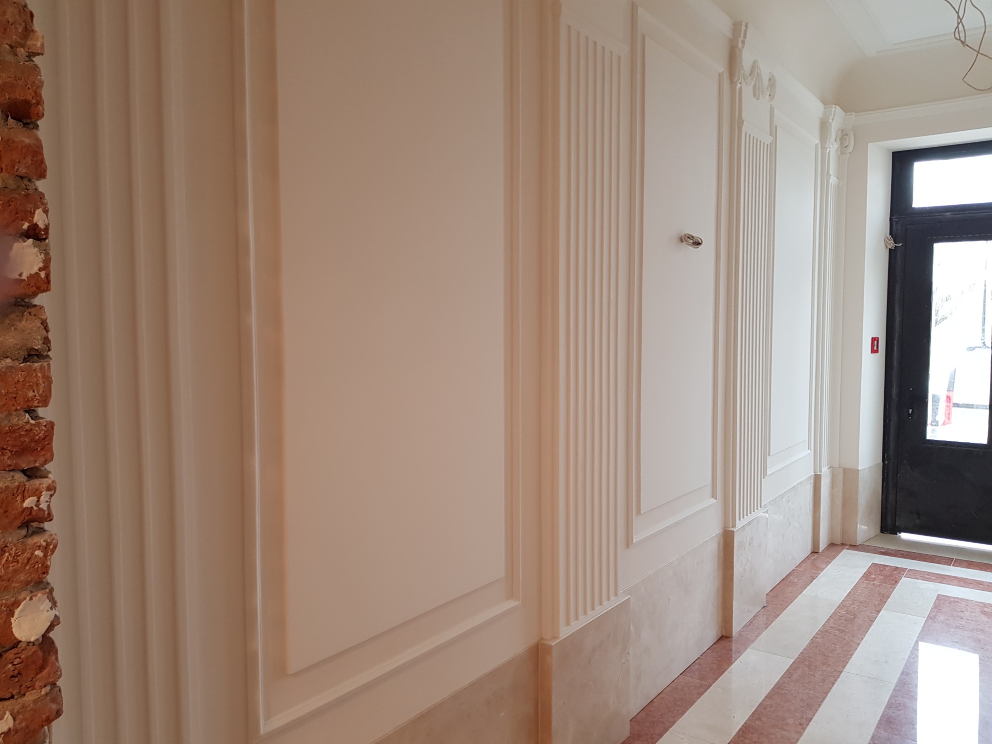 Korytarz po remoncie z dekoracyjnymi panelami ściennymi w odcieniach bieli i beżu, z fragmentem surowej cegły po lewej stronie i marmurową podłogą w paski.