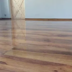 Błyszcząca, nowo położona podłoga laminowana o wzorze drewna, widoczna drewniana listwa przypodłogowa i fragment drzwi z jasnego drewna.