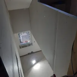 Wnętrze poddasza w trakcie wykańczania, widok z dołu na okno dachowe i zamontowane płyty gipsowo-kartonowe na konstrukcji metalowej, widoczne ślady po szpachlowaniu, oświetlenie robocze i fragment...