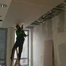 Pracownik na drabinie montuje płytę gipsową do metalowego stelażu sufitu podwieszanego w pomieszczeniu z oknami i częściowo wykończonymi ścianami.