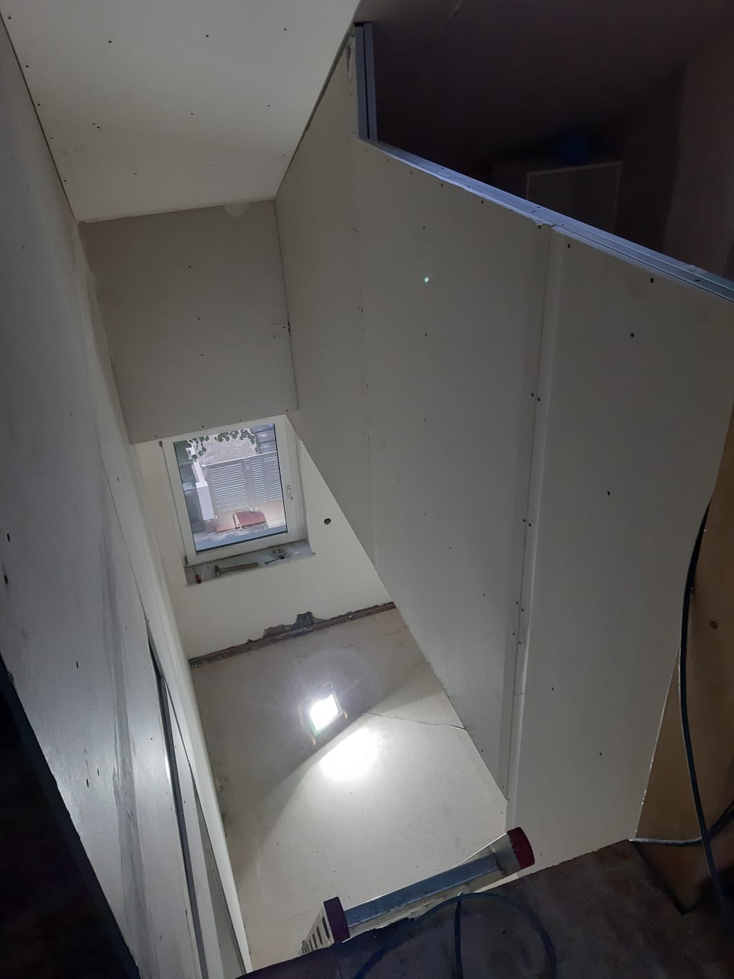 Sufit poddasza w trakcie wykańczania płytami gipsowo-kartonowymi, widoczne metalowe profile i oświetlenie robocze, okno w tle.