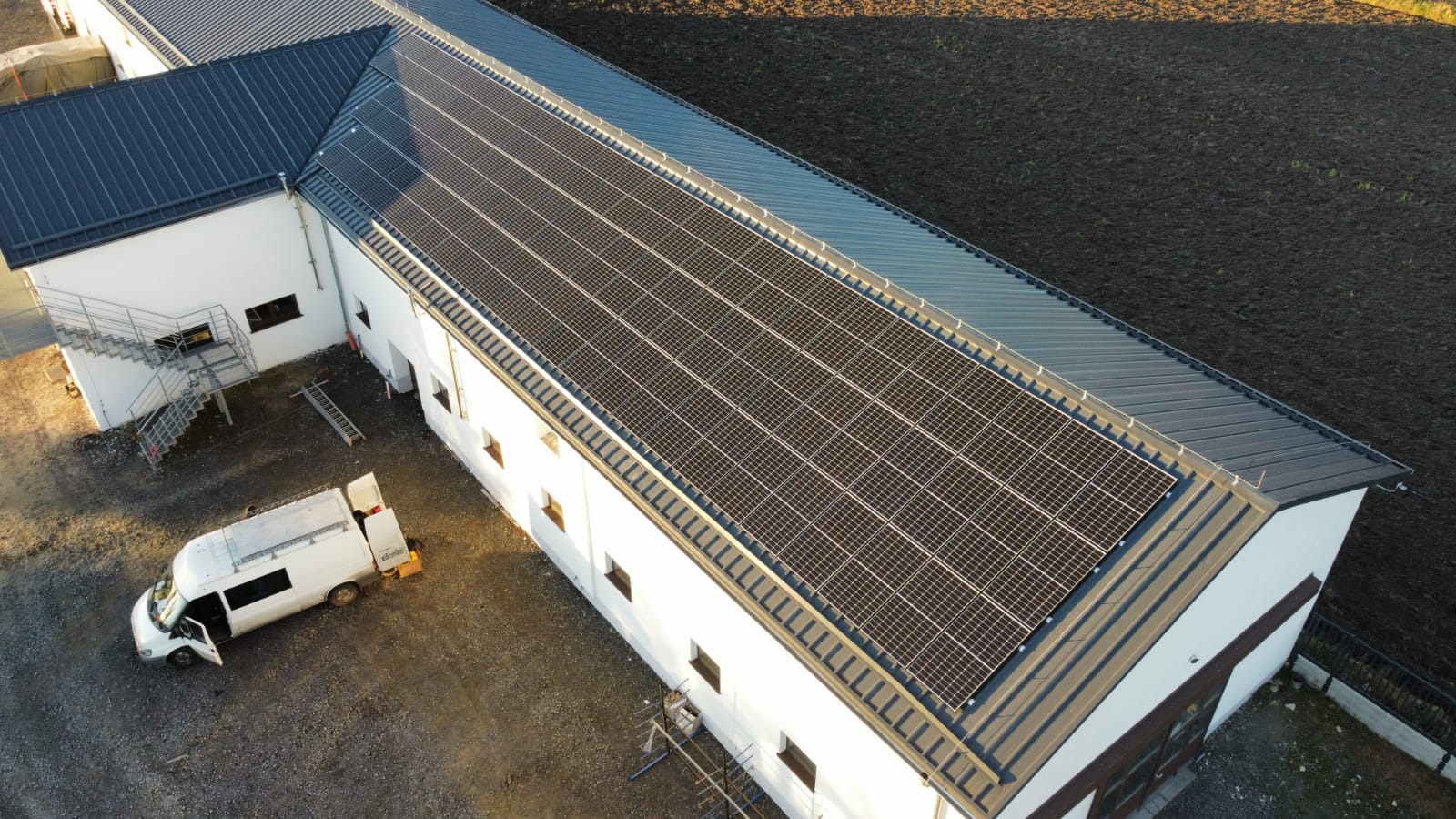 Widok z góry na budynek z panelami słonecznymi na dachu, obok zaparkowany biały bus serwisowy z otwartymi drzwiami, na zewnątrz budynku metalowe schody i drabina.