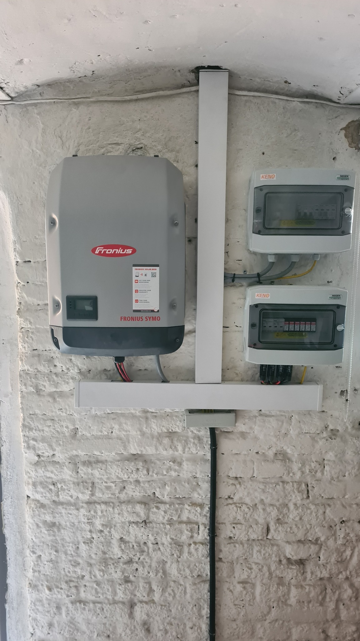 Szary inwerter Fronius Symo zamontowany na białej, chropowatej ścianie z cegły, widoczne również dwie szare skrzynki z bezpiecznikami KENO, całość połączona białymi profilami.