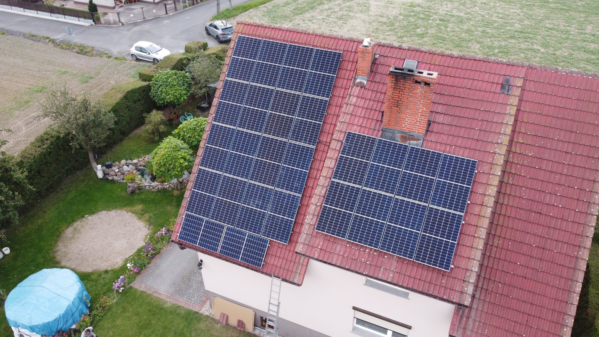 Widok z drona na dom z czerwoną dachówką i dwoma zestawami paneli słonecznych, z drabinką opartą o ścianę, ogród z altaną i dwoma samochodami w tle.