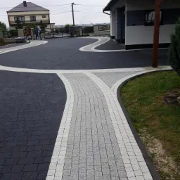 Nowoczesny podjazd z kostki brukowej w odcieniach szarości i czerni, z dekoracyjnymi liniami, prowadzący do domu z garażem i ogrodzeniem w tle.
