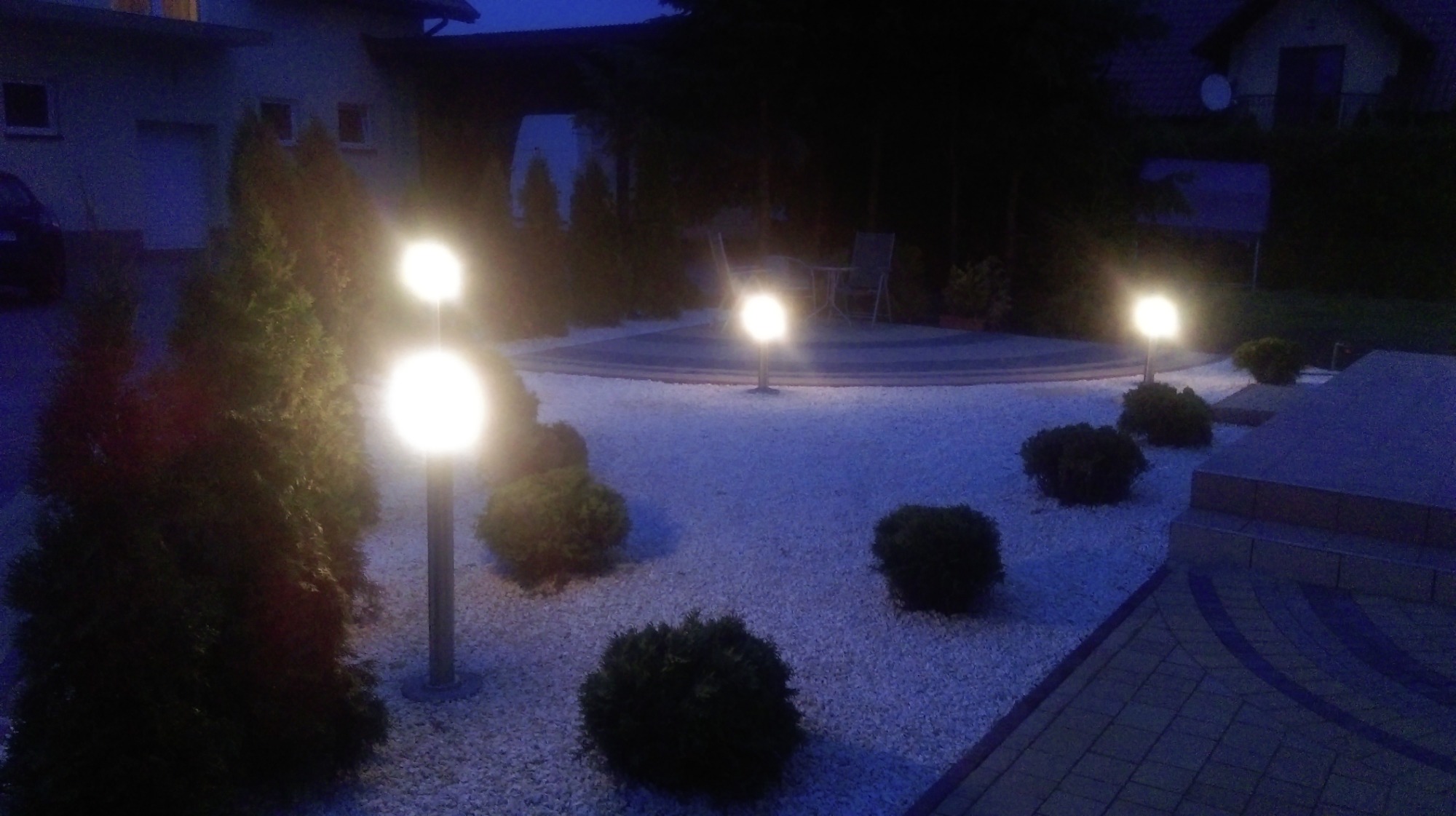 Oświetlony ogród nocą z okrągłym tarasem, wysypany białym żwirem, z kulistymi lampami i niskimi krzewami.