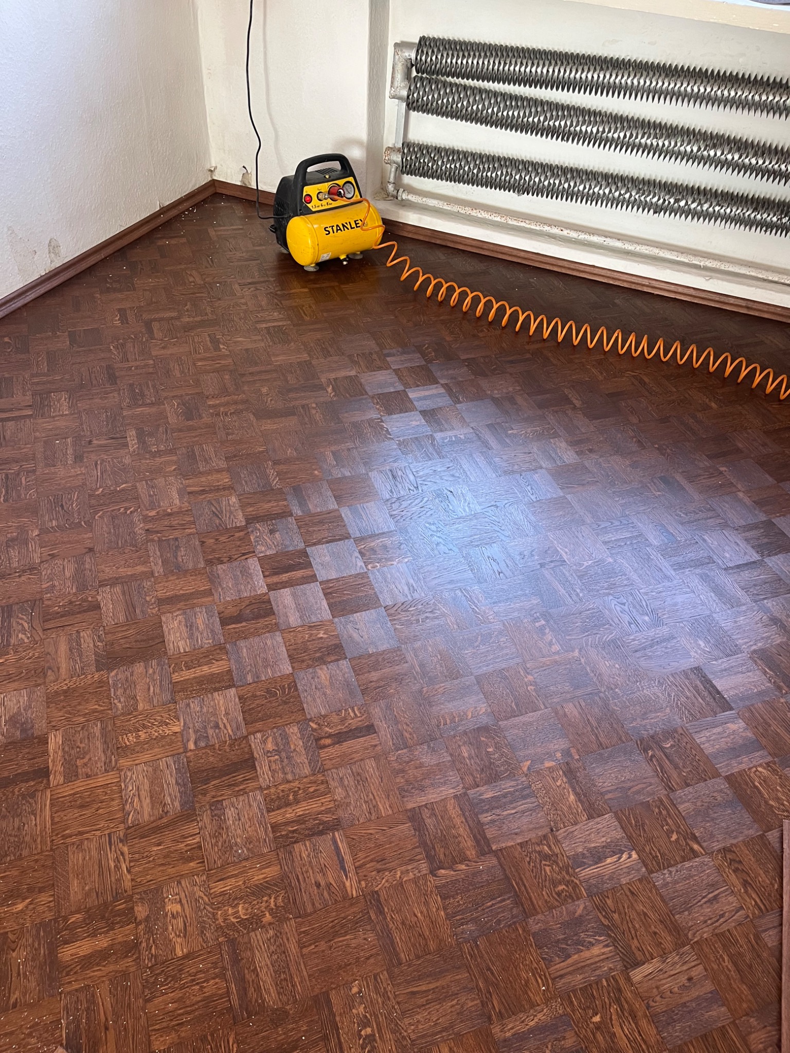 Parkiet w kwadraty po obróbce, widoczny żółty kompresor Stanley z pomarańczowym wężem pneumatycznym i grzejnik żeberkowy.