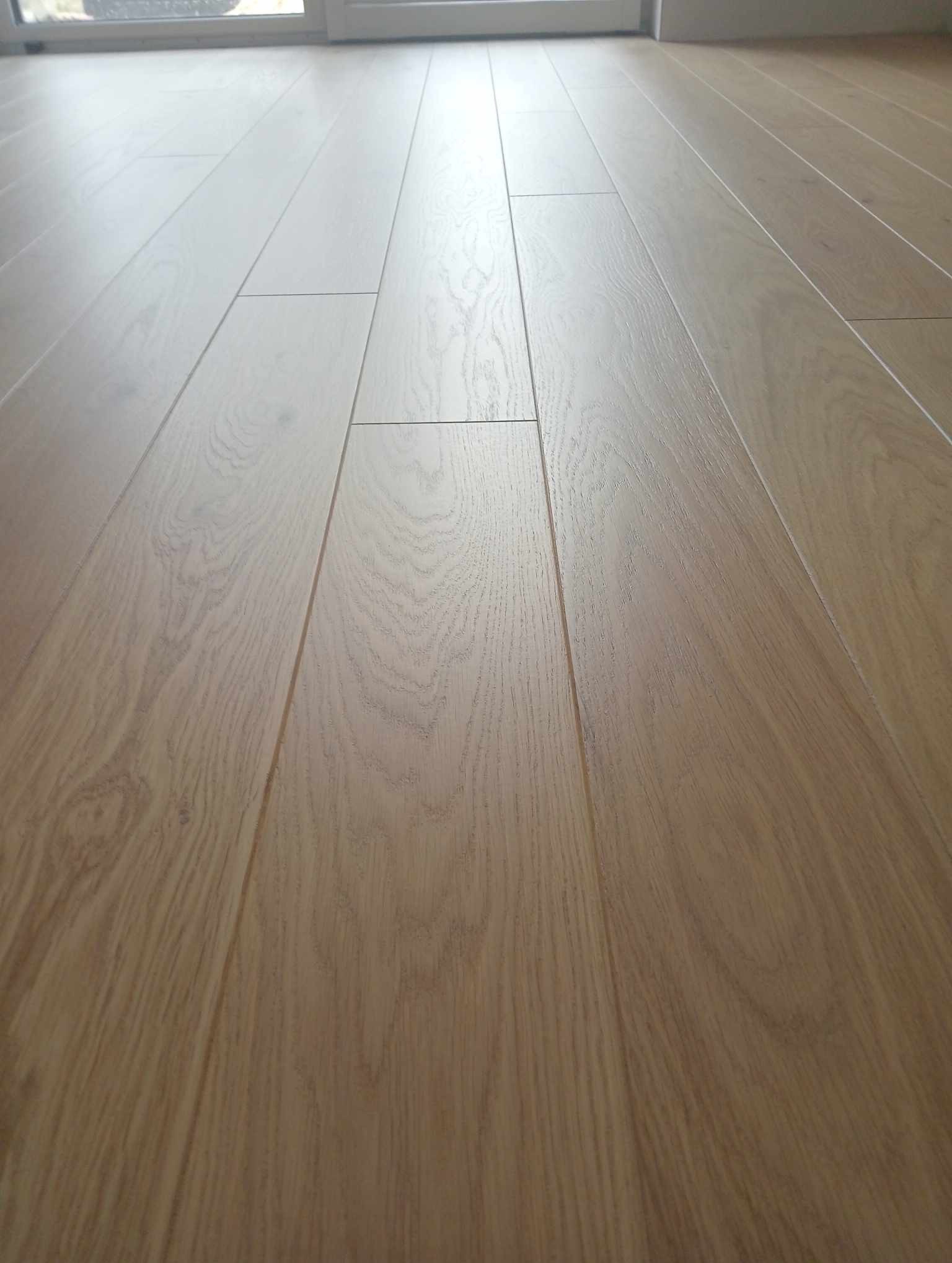 Jasny, dębowy parkiet ułożony w regularny wzór, widoczny w perspektywie z niskiego kąta, z delikatnym połyskiem i widocznymi słojami drewna.