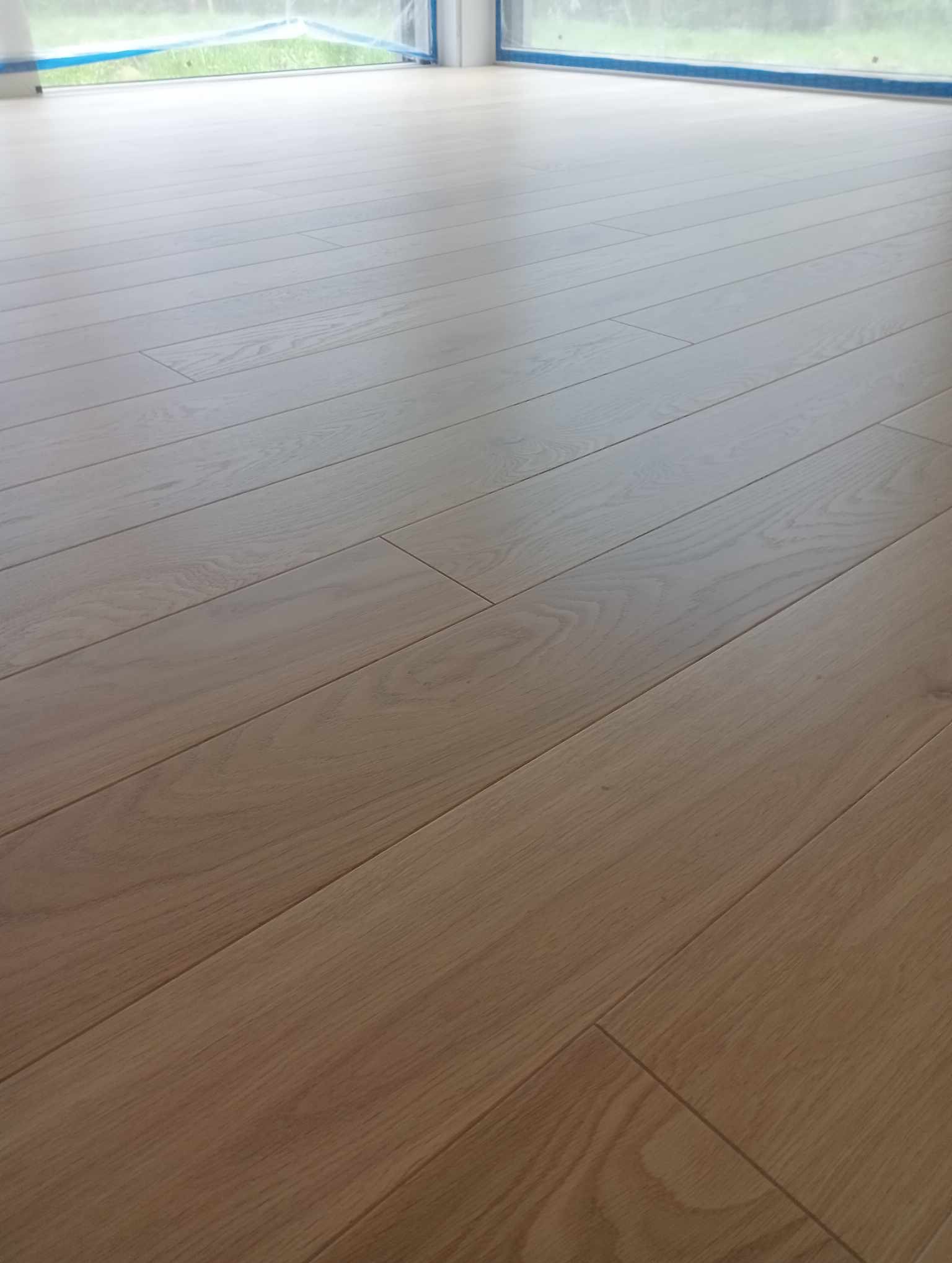 Jasna, drewniana podłoga ułożona w jodełkę, widok z niskiego kąta, okno zabezpieczone folią malarską w tle.