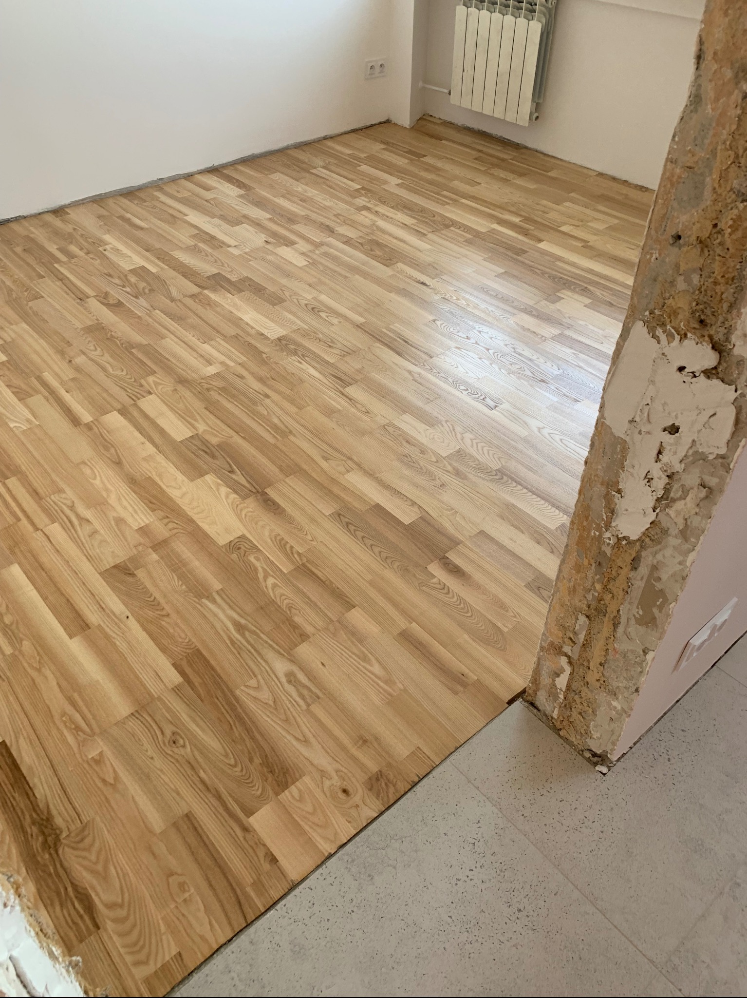 Ułożony parkiet laminowany w jasnym odcieniu drewna, widoczny fragment surowej ściany z resztkami tynku oraz grzejnik w tle.