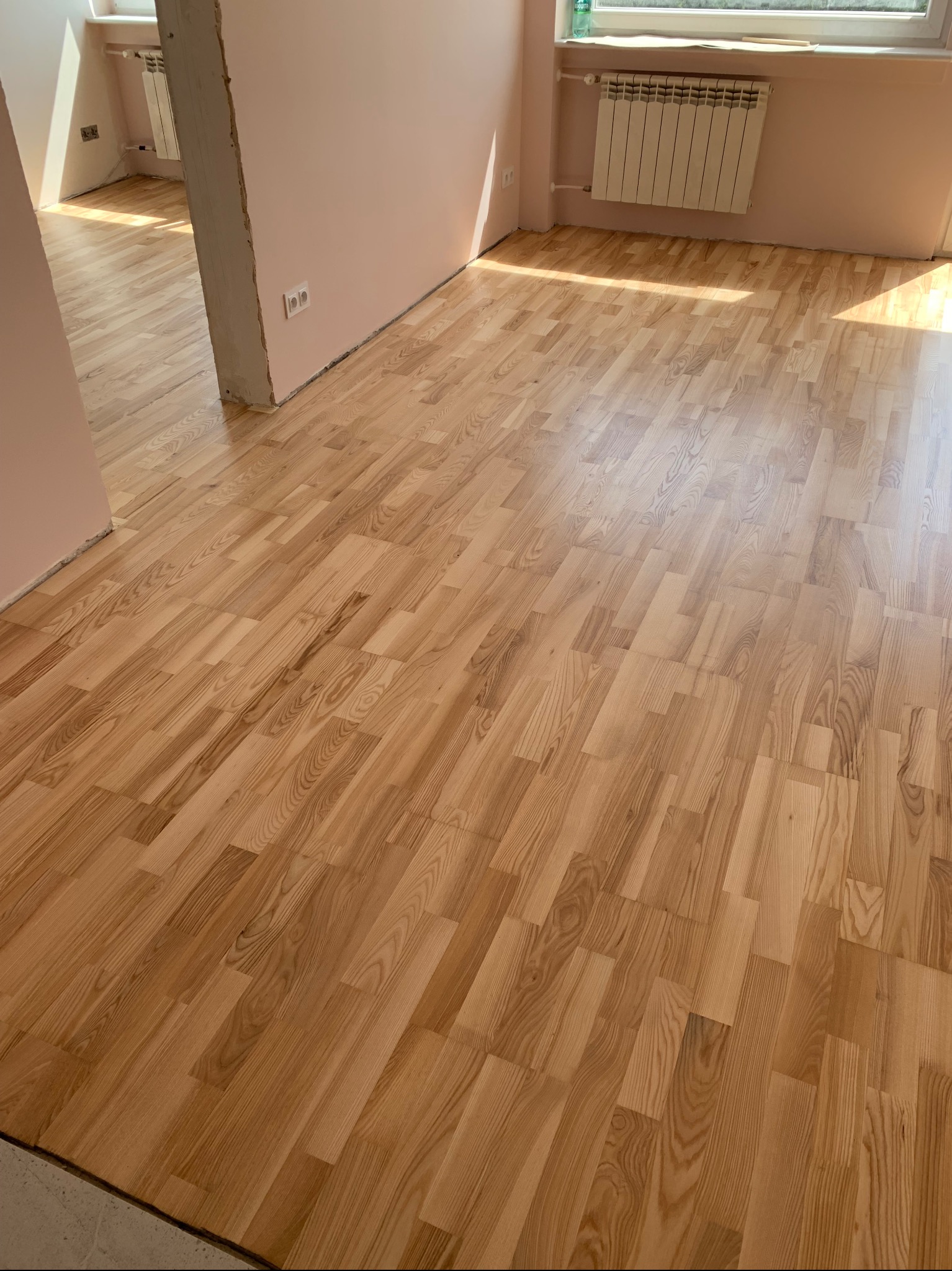 Świeżo ułożona, jasna podłoga parkietowa w jodełkę klasyczną, w pokoju z różowymi ścianami i grzejnikiem pod oknem, widok z góry.