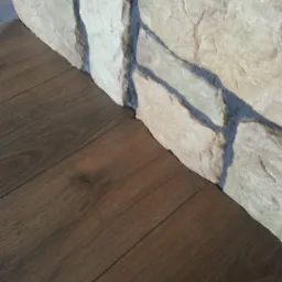 Ciemnobrązowa podłoga z desek sąsiadująca z kamienną ścianą o nieregularnej fakturze i jasnych odcieniach beżu i szarości.